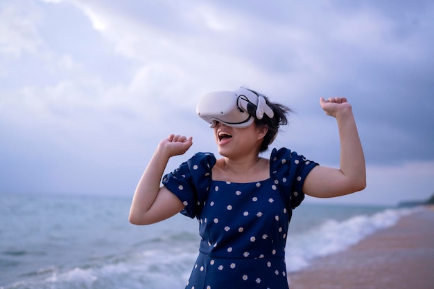 Feliz, tierna, alegre, atractiva, mujer adulta asiática, ropa casual con gafas de tecnología VR de pie en la playa y ajustando el dispositivo para ver video digital virtual de simulación 3D en la naturaleza real