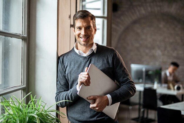 Feliz trabajador independiente con laptop mirando a la cámara mientras está de pie junto a la ventana de la oficina