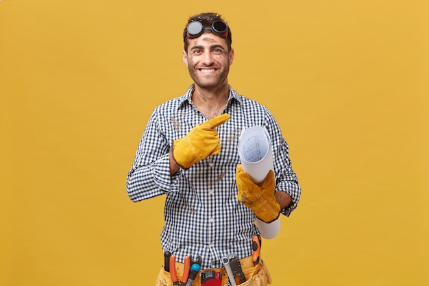 Feliz trabajador de mantenimiento o reparador con la cara sucia con gafas protectoras, guantes y cinturón con instrumentos que sostienen el plano de pie contra la pared amarilla en blanco apuntando a copyspace