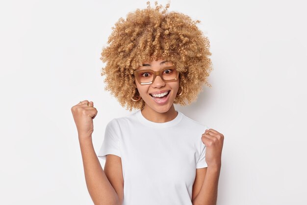 Feliz triunfante joven con cabello rizado aprieta los puños celebra el éxito feliz de lograr metas vestida con camiseta casual aislada sobre fondo blanco. Modelo femenino ambicioso y exitoso