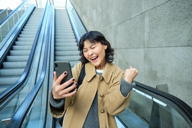 Feliz triunfante niña asiática se ve asombrada y alegre sostiene que el teléfono inteligente baja las escaleras mecánicas con por favor