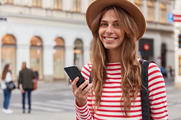 Feliz turista usa información del blog de viajes, sostiene un teléfono inteligente, camina por las calles de la ciudad, usa un elegante sombrero y un jersey a rayas