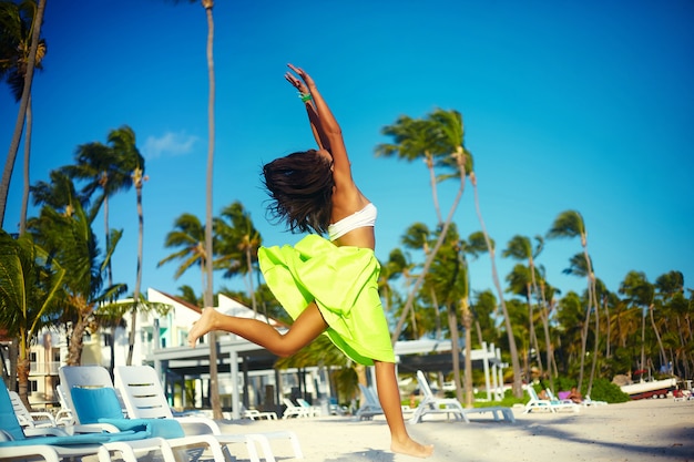 Feliz urbano moderno joven elegante mujer modelo mujer en tela moderna brillante en falda colorida verde al aire libre en la playa de verano saltando detrás del cielo azul