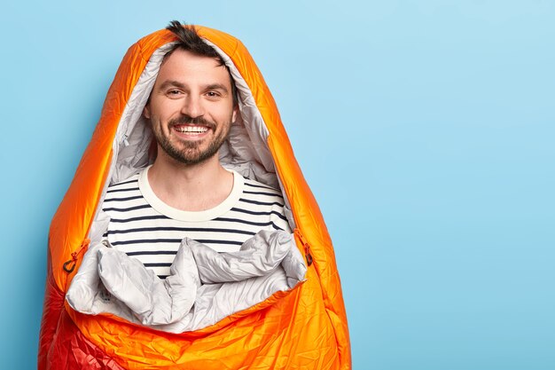 Feliz vacacionista sin afeitar pasa las vacaciones cerca de las montañas en el campamento, duerme en un saco de dormir, sonríe positivamente, vestido con un suéter a rayas, posa contra la pared azul