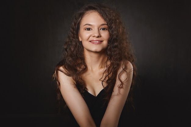 Feminidad, belleza y lenguaje corporal. Disparo aislado de linda chica guapa con cabello oscuro rizado y maquillaje sentado en el interior en postura cerrada