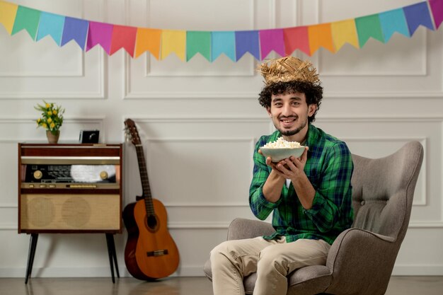 Festa junina lindo joven con sombrero de paja con radio retro y bandera colorida sosteniendo un tazón de palomitas de maíz