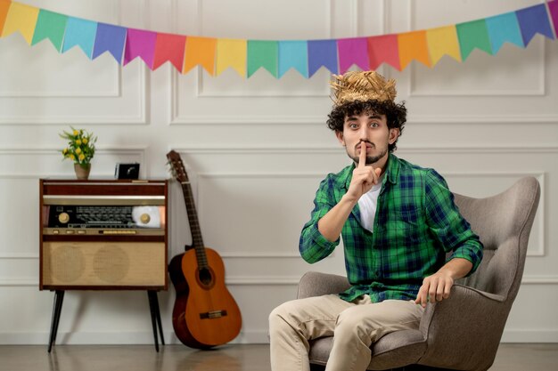 Festa junina lindo joven con sombrero de paja con radio retro y banderas coloridas que muestran gesto de silencio