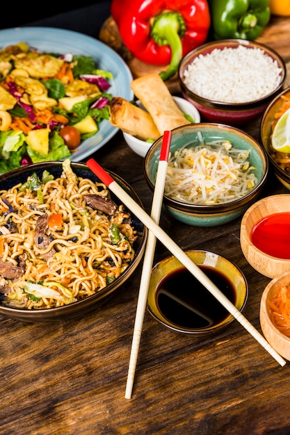 Fideos tailandeses; ensalada; rollitos de primavera; arroz; brotes de alubias; Salsas con palillos en mesa de madera.