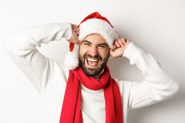 Fiesta de año nuevo y concepto de vacaciones de invierno. Primer plano de hombre barbudo alegre celebrando la Navidad, sonriendo y vistiendo gorro de Papá Noel, fondo blanco.