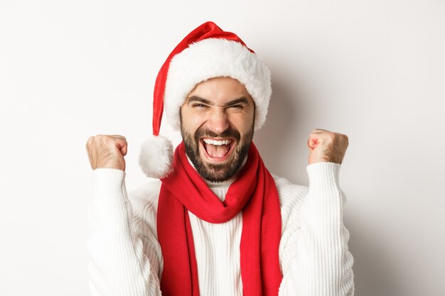 Fiesta de año nuevo y concepto de vacaciones de invierno. Primer plano del hombre emocionado en el regocijo del sombrero de Santa, ganando o celebrando en el sombrero de Santa, de pie contra el fondo blanco