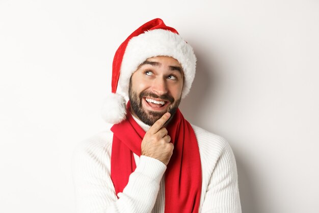 Fiesta de año nuevo y concepto de vacaciones de invierno. Primer plano de hombre guapo con barba mirando pensativo, planificación de la lista de regalos de Navidad en el gorro de Papá Noel, fondo blanco.