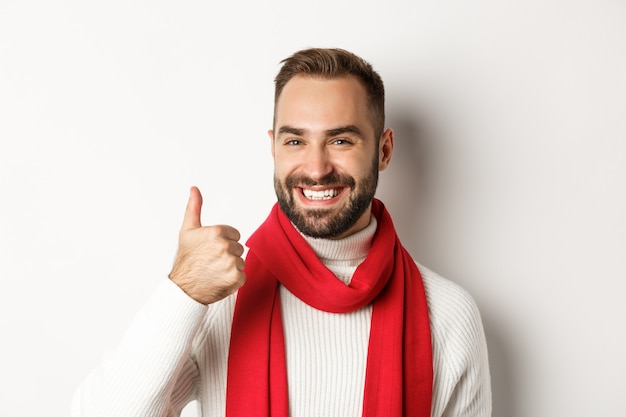 Fiesta de año nuevo y concepto de vacaciones de invierno. Primer plano de hombre guapo satisfecho mostrando el pulgar hacia arriba, deseando feliz Navidad, de pie sobre fondo blanco.