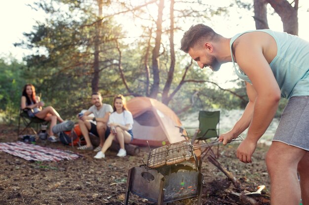 Fiesta, campamento de grupo de hombres y mujeres en el bosque. Se relajan, cantan una canción y cocinan barbacoa contra la hierba verde. Las vacaciones, verano, aventura, estilo de vida, concepto de picnic.