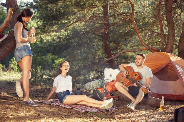 Fiesta, campamento de grupo de hombres y mujeres en el bosque. Se relajan, cantan una canción contra la hierba verde. Las vacaciones, verano, aventura, estilo de vida, concepto de picnic.