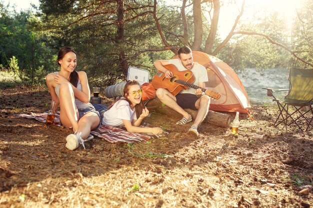 Fiesta, campamento de grupo de hombres y mujeres en el bosque. Se relajan, cantan una canción contra la hierba verde. Las vacaciones, verano, aventura, estilo de vida, concepto de picnic.