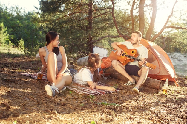 Fiesta, campamento de grupo de hombres y mujeres en el bosque. Se relajan, cantan una canción contra la hierba verde. Las vacaciones, verano, aventura, estilo de vida, concepto de picnic.