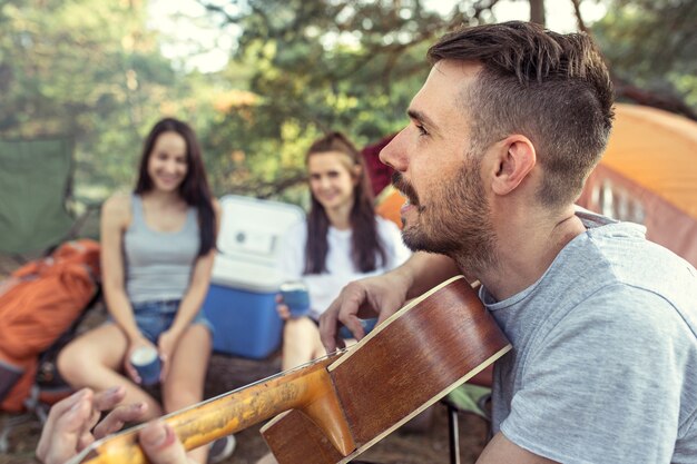 Fiesta, campamento de grupo de hombres y mujeres en el bosque. Se relajan, cantan una canción contra la hierba verde. Las vacaciones, verano, aventura, estilo de vida, concepto de picnic.