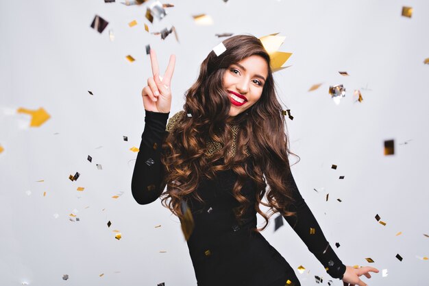 Fiesta de cumpleaños, carnaval de año nuevo. Joven sonriente celebrando un evento brillante, viste un elegante vestido negro de moda y una corona amarilla. Confeti espumoso, divertirse, bailar.