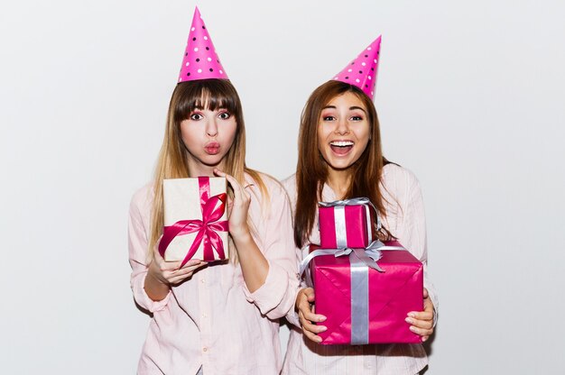 Fiesta de cumpleaños de pijamas. Amigos divirtiéndose y sosteniendo cajas de regalo. Cara de sorpresa, emociones emocionadas. Chicas con sombreros de fiesta de utilería. Interior.