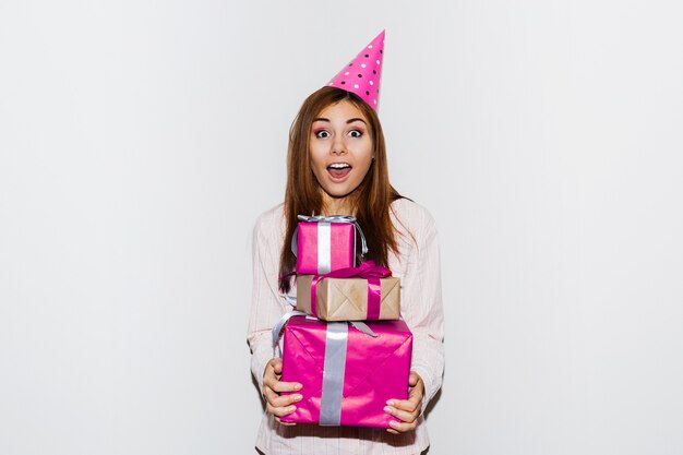 Fiesta de cumpleaños de pijamas. Linda chica con cara de sorpresa sosteniendo cajas de regalo, con sombreros de fiesta de utilería. Retrato flash.