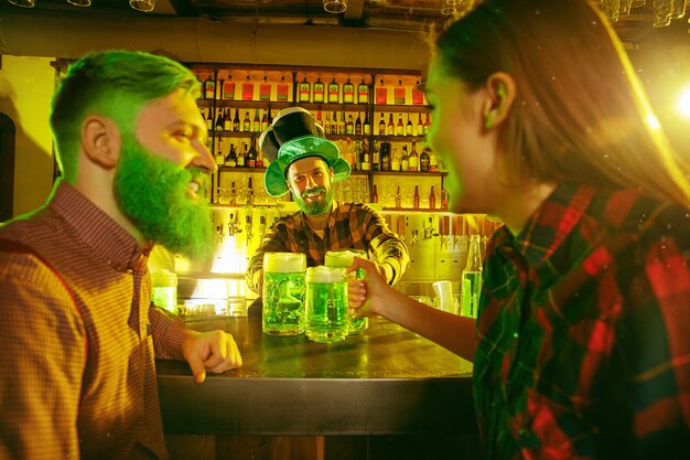 Fiesta del día de San Patricio. Amigos felices están celebrando y bebiendo cerveza verde. Hombres y mujeres jóvenes con sombreros verdes. Interior del pub.