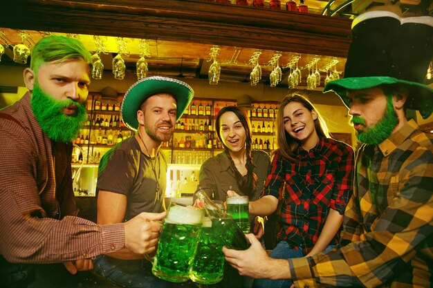 Fiesta del día de San Patricio. Amigos felices están celebrando y bebiendo cerveza verde. Hombres y mujeres jóvenes con sombreros verdes. Interior del pub.