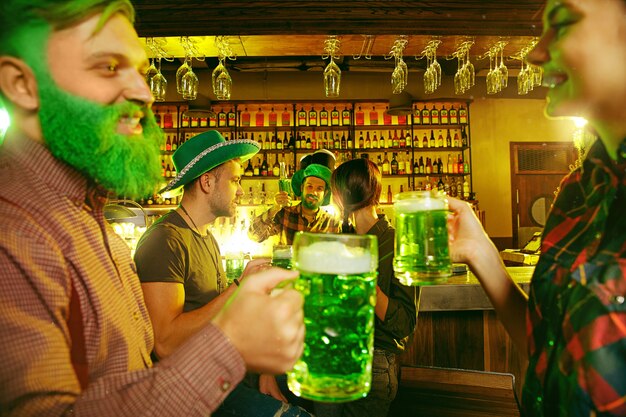 Fiesta del día de San Patricio. Amigos felices están celebrando y bebiendo cerveza verde. Hombres y mujeres jóvenes con sombreros verdes. Interior del pub.