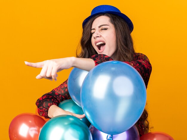 Fiesta joven mujer con sombrero de fiesta de pie detrás de globos apuntando al lado gritando con los ojos cerrados aislado en la pared naranja