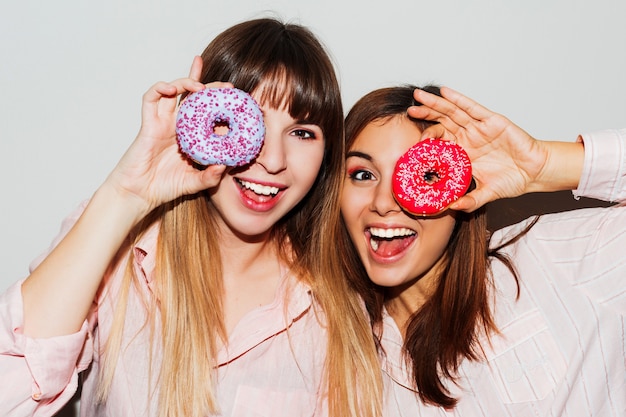 Fiesta de pijamas en casa. Primer plano retrato flash de dos divertidas mujeres blancas posando con donas. Cara de sorpresa.