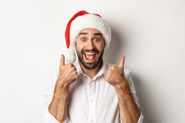 Fiesta, vacaciones de invierno y concepto de celebración. Hombre disfrutando de la Navidad, vistiendo gorro de Papá Noel y mostrando el pulgar hacia arriba con cara emocionada
