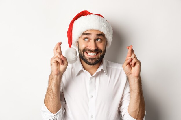 Fiesta, vacaciones de invierno y concepto de celebración. Hombre feliz con gorro de Papá Noel haciendo deseos de Navidad, cruzar los dedos para la buena suerte y sonriendo emocionado