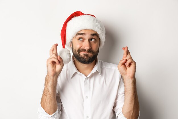 Fiesta, vacaciones de invierno y concepto de celebración. Hombre feliz con gorro de Papá Noel que desea Navidad, cruza los dedos para la buena suerte y parece esperanzado en la esquina superior izquierda.