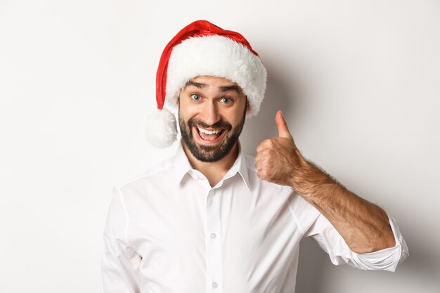 Fiesta, vacaciones de invierno y concepto de celebración. Primer plano de hombre barbudo satisfecho con sombrero de santa mostrando el pulgar hacia arriba, aprobar y como algo bueno