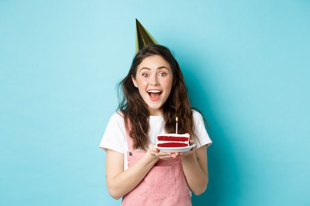 Fiestas y celebraciones. Feliz cumpleañera con sombrero de fiesta sosteniendo pastel de cumpleaños y sonriendo, pidiendo deseos en la vela encendida, de pie contra el fondo azul.