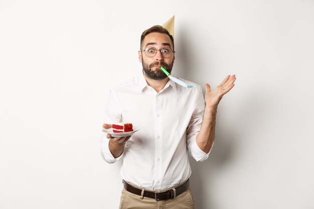 Fiestas y celebraciones. Hombre alegre disfrutando de cumpleaños, soplando el silbato de la fiesta y sosteniendo el pastel de cumpleaños