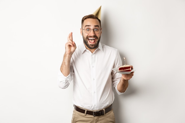 Fiestas y celebraciones. Hombre emocionado que tiene una fiesta de cumpleaños, pide un deseo en el b-day cake y cruza los dedos para la buena suerte, de pie