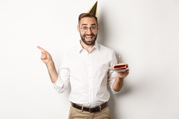 Fiestas y celebraciones. Hombre feliz disfrutando de la fiesta de cumpleaños, sosteniendo el pastel de cumpleaños y señalando con el dedo a la izquierda en la promoción, de pie