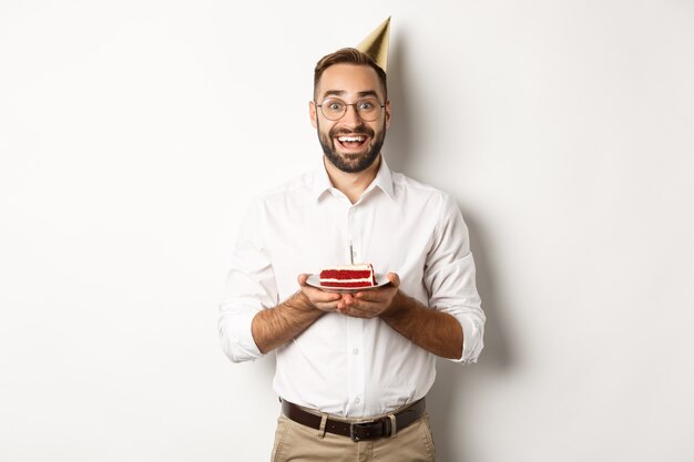 Fiestas y celebraciones. Hombre feliz con fiesta de cumpleaños, pidiendo deseos en b-day cake y sonriendo, de pie