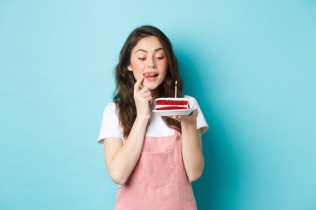 Fiestas y celebraciones. Linda cumpleañera mirando sabroso pastel con la tentación de morderlo, de pie soñadora contra el fondo azul.