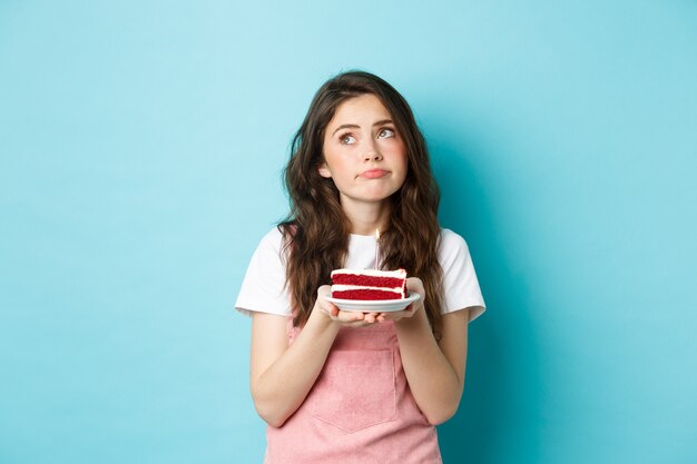 Fiestas y celebraciones. Mujer triste y sombría que sostiene la torta de cumpleaños, mirando a otro lado con cara pensativa molesta, de pie sobre fondo azul.