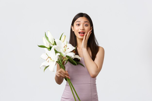 Fiestas y eventos, concepto de celebración. Chica asiática feliz sorprendida en traje de noche, recibe ramo de flores de admirador secreto, jadeando asombrado y conmovido, sosteniendo lirios, fondo blanco.