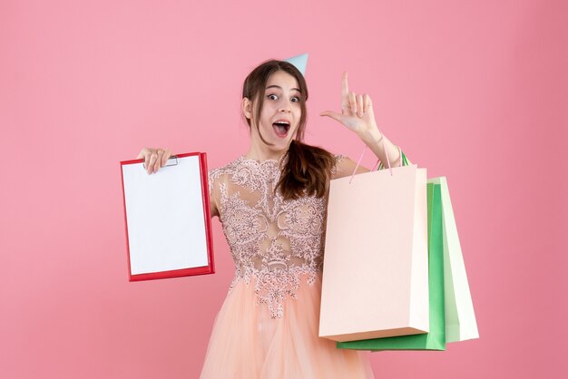 fiestera con gorra de fiesta sosteniendo documentos y bolsas de compras apuntando con el dedo hacia arriba en rosa