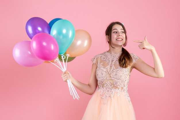 fiestera con gorra de fiesta sosteniendo globos apuntando a su sonrisa en rosa