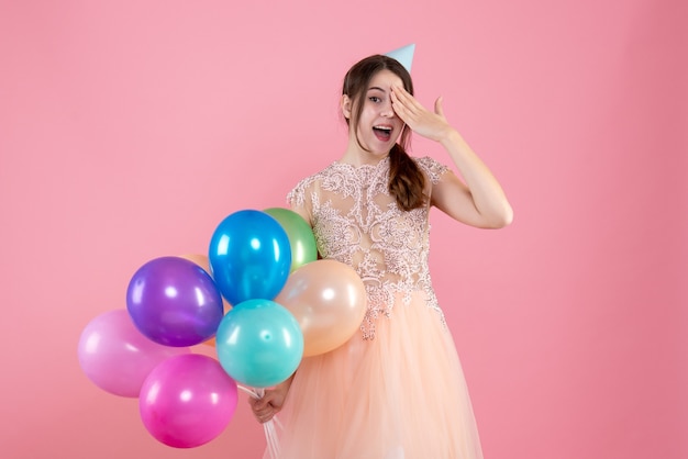 Foto gratuita fiestera con gorra de fiesta sosteniendo globos poniendo la mano en el ojo en rosa