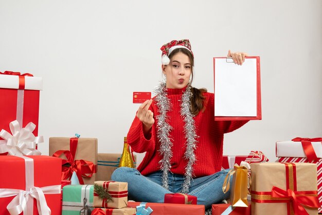 Foto gratuita fiestera con gorro de papá noel con tarjeta y documentos sentados alrededor presenta en blanco