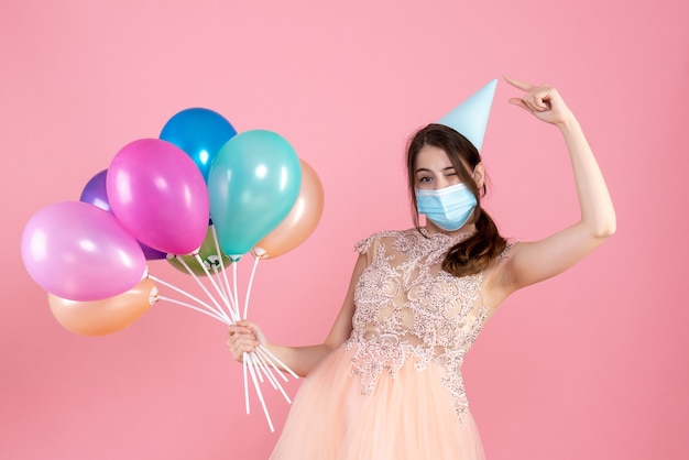 fiestera con máscara médica apuntando a su gorra de fiesta sosteniendo globos de colores en rosa