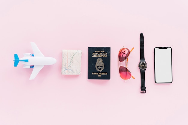 Fila de avión de juguete; mapa plegado; pasaporte; Gafas de sol; Reloj de pulsera y teléfono inteligente sobre fondo rosa.