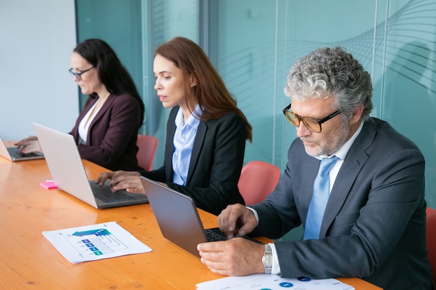 Fila de gente de negocios enfocada que trabaja en computadoras en una mesa con gráficos de papel