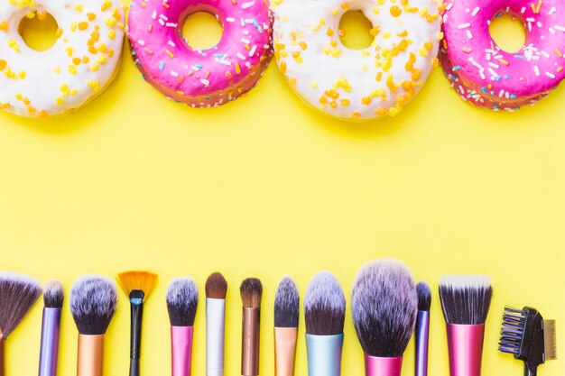 Fila de pinceles de maquillaje y donas sobre fondo amarillo