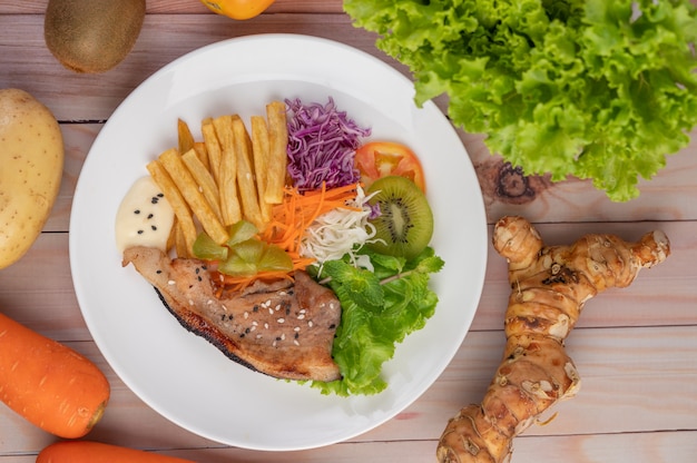 Filete de pescado con papas fritas, kiwi, lechuga, zanahorias, tomates y repollo en un plato blanco.